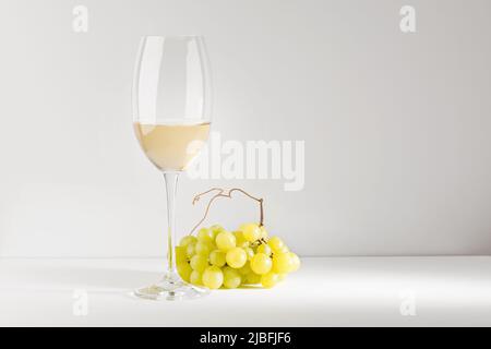 Vin blanc dans un gobelet en verre et un bouquet de raisins sur une table blanche. Arrière-plan clair. Banque D'Images