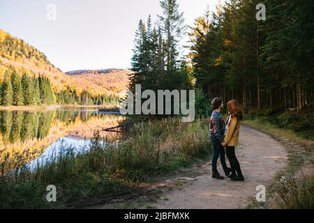 Vue latérale d'un jeune couple romantique en vêtements élégants embrassant et admirant le paysage d'automne pittoresque du lac calme entouré d'une forêt luxuriante Banque D'Images