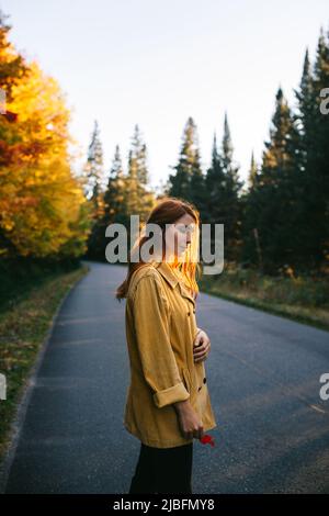 Vue latérale de la jeune femme pensive touriste avec de longs cheveux bruns debout avec les yeux fermés dans des vêtements décontractés tenant la feuille d'érable doré à la main sur l'asphal Banque D'Images