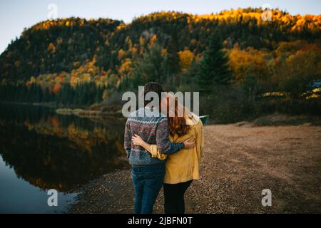 Vue arrière d'un couple de voyage romantique et méconnaissable avec des vêtements élégants embrassant et admirant le paysage d'automne pittoresque d'un lac calme entouré de lus Banque D'Images