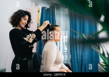 Vue latérale d'un coiffeur positif dans des vêtements décontractés souriant et ajustant la queue de cheval du client hispanique heureux pendant le travail dans le salon Banque D'Images