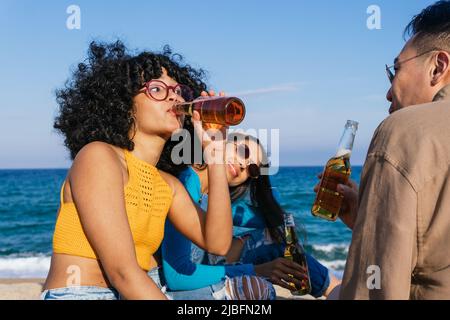 Femme afro-américaine vêtie de vêtements décontractés aux cheveux bouclés en sirotant de la bière à la bouteille tout en passant du temps avec divers amis sur la plage près de la mer Banque D'Images