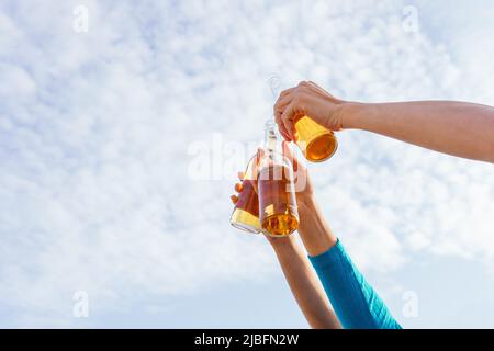 De dessous anonyme des mains de culture d'homme et de femmes élevant des bouteilles de bière tout en se refroidissant sur la plage contre le ciel bleu le jour de week-end d'été Banque D'Images