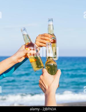 Des mains anonymes d'hommes et de femmes qui élèvent des bouteilles de bière tout en se refroidissant sur la plage contre l'eau de mer et le ciel bleu le jour du week-end d'été Banque D'Images