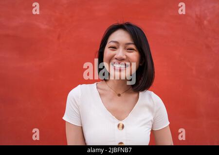 Jeune femme asiatique joyeuse et sûre d'elle-même avec des cheveux sombres dans des vêtements décontractés souriant heureux et regardant l'appareil photo sur fond rouge Banque D'Images