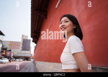 Vue latérale jeune femme asiatique joyeuse millénaire avec des cheveux sombres dans des vêtements décontractés souriant heureux et regardant loin sur fond rouge Banque D'Images