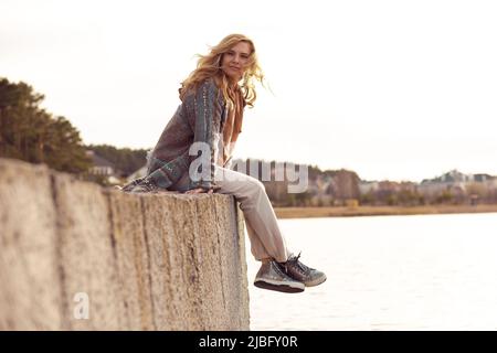 Portrait d'une jeune femme souriante avec de longs cheveux ondulés et justes assis avec des jambes croisées sur le bord de la rive de rivière en béton. Banque D'Images