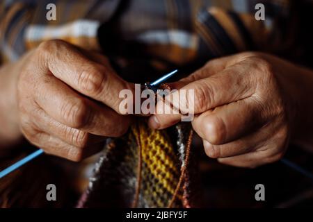 Gros plan des mains d'une femme âgée méconnue portant une chemise à carreaux, tricoter une écharpe colorée avec des aiguilles en métal. Banque D'Images