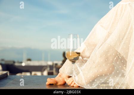 Photo courte des jambes pieds nus d'une femme vêtue d'une longue robe d'été blanche et transparente, avec un verre de champagne. Banque D'Images