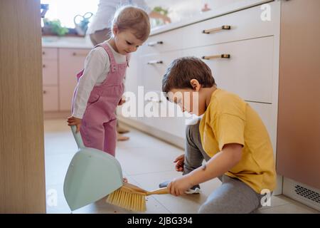 Un petit garçon et une petite fille qui aident à nettoyer la maison à l'aide d'une casserole et d'une brosse qui balaient la saleté du sol. Banque D'Images