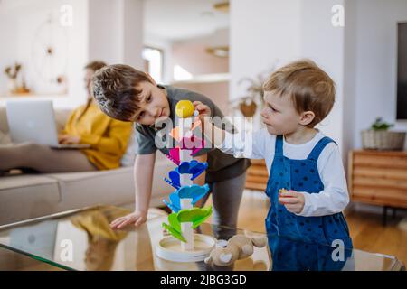 Petits frères et sœurs jouant avec le marbre en bois montessori dans la salle de séjour, leur mère est assise sur un canapé avec ordinateur portable. Banque D'Images