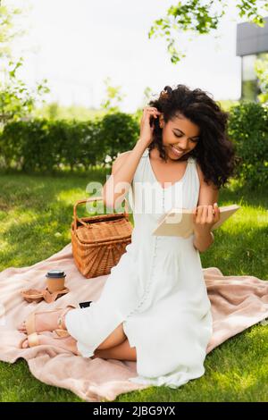 joyeuse femme afro-américaine en robe lisant le roman et assis sur une couverture dans le parc Banque D'Images