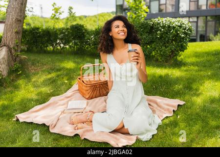 joyeuse femme afro-américaine en robe tenant une tasse de papier et assise sur une couverture près du panier de pique-nique Banque D'Images