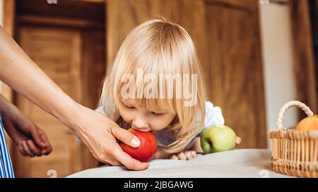 Une petite fille mord une pomme qui est tenue par une maman ou une gardienne assise à la table Banque D'Images