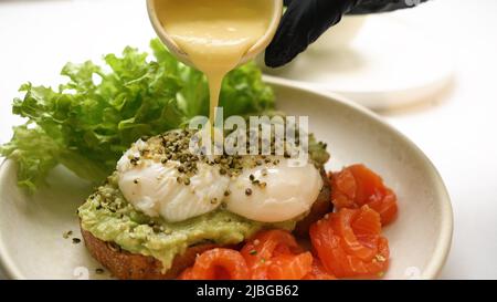 Le chef verse de la sauce sur des toasts au guacamole et des œufs pochés parsemés de graines. Truite et feuilles de salade fraîches servies sur le côté du toast en gros plan Banque D'Images