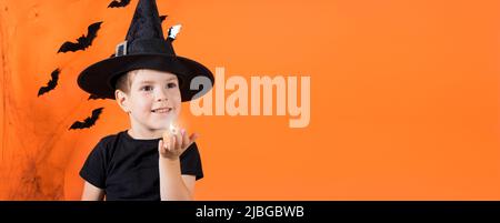 Un garçon en costume de sorcière dans un chapeau noir tient une petite lampe de poche à citrouille Jack. Sur un fond orange. Halloween enfants. Emplacement de la bannière pour Banque D'Images