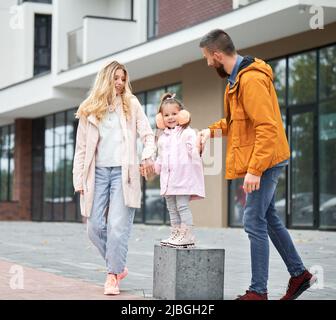 Pleine longueur d'homme barbu et de femme blonde jouant avec une petite fille dans la rue dans le nouveau quartier urbain. Une famille heureuse et aimante avec un enfant qui s'amuse à l'extérieur. Banque D'Images