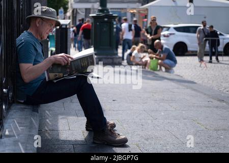 Un homme plus âgé dans un chapeau de lin est assis sur un mur sur la Markt ensoleillée de Maastricht et lit le journal. Une jeune famille et des stands de marché flous dans le dos Banque D'Images