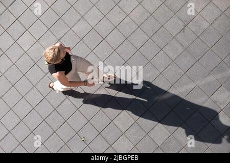 Une jeune femme non identifiée aux cheveux blonds et au téléphone à la main marche sur un trottoir à Maastricht. Vue d'en haut, ombre longue Banque D'Images