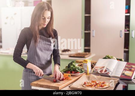 Les jeunes femmes cuisent des ingrédients pour la pizza sur la planche à découper en suivant la recette écrite dans un livre debout dans la cuisine. Banque D'Images