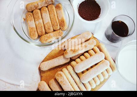 Pose à plat. Ingrédients pour préparer du tiramisu italien traditionnel sur une table de cuisine - biscuits savoiardi sur une planche à bois, café expresso sucré en g Banque D'Images