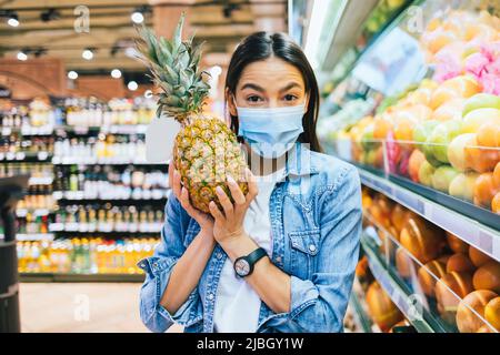 Jeune femme gaie dans un masque de protection tenant l'ananas tout en se tenant dans un supermarché faisant des courses Banque D'Images