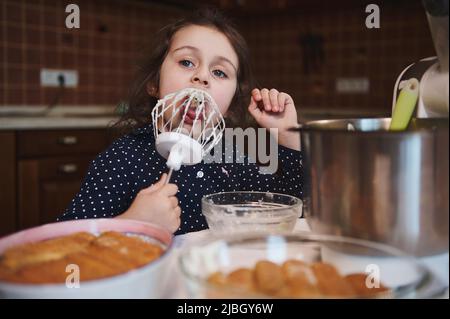 Portrait d'une adorable petite fille caucasienne de 5 ans léchant les restes de crème fouettée douce sur un fouet debout à la table de cuisine avec tiramisu flou Banque D'Images