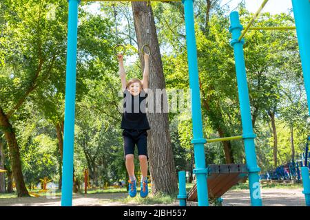 le garçon entre pour le sport dans le parc il est suspendu sur les anneaux de gymnastique et essaie de se tirer vers le haut Banque D'Images