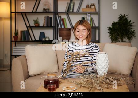 Jeune femme vietnamienne souriante et talentueuse qui boit du thé et organise un bouquet de fleurs séchées Banque D'Images