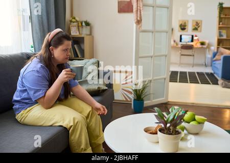 Jeune femme sérieuse à la maison porte mug avec thé chaud tout en s'asseyant sur un canapé confortable et doux et prenant le petit déjeuner Banque D'Images