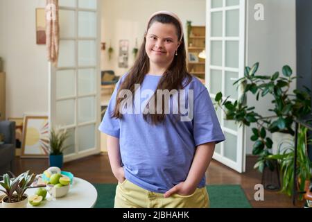 Fille gaie avec le syndrome de Down portant un t-shirt bleu et un pantalon en coton jaune debout devant l'appareil photo dans le salon spacieux Banque D'Images