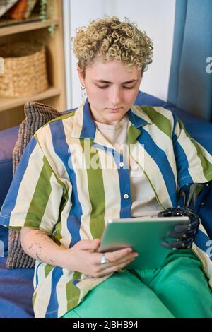 Jeune femme sérieuse avec prothèse de bras regardant l'écran de tablette tout en étant assise sur un canapé confortable dans le salon et travaillant dans le filet Banque D'Images