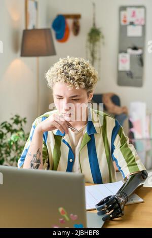 Jeune femme sérieuse avec bras myoélectrique regardant l'écran d'ordinateur portable tout en travaillant sur un nouveau projet d'entreprise par bureau dans le bureau à domicile Banque D'Images