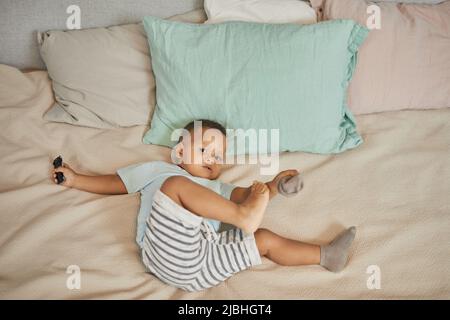 Vue en grand angle d'un adorable tout-petit noir posé sur un lit dans des draps de couleur pastel et jouant avec des chaussettes regardant l'appareil photo Banque D'Images