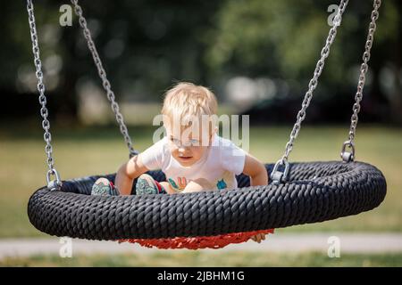 Petit garçon blond jouant avec joie sur un grand balançoire de nid à l'aire de jeux de la ville. Siège rond noir et rouge pour balancer les enfants. Banque D'Images