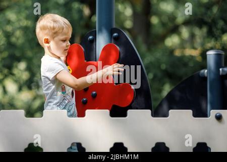 Un adorable garçon sur un terrain de jeu extérieur, un gouvernail de bateau rouge, une clôture décorative, un fond de feuillage vert. Banque D'Images