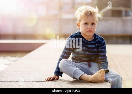 Petit garçon triste assis près de la fontaine un jour ensoleillé, l'enfant regarde la caméra photo, il y a de l'espace libre pour le texte dans l'image. Loisirs d'été Banque D'Images