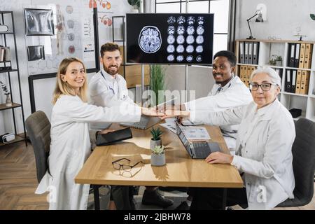 Portrait d'un groupe multiracial de médecins heureux tenant les mains ensemble tout en étant assis à table à l'hôpital. Vue d'ensemble des experts en soins de santé se réjouissant de la réussite de la réunion et des bonnes nouvelles. Banque D'Images