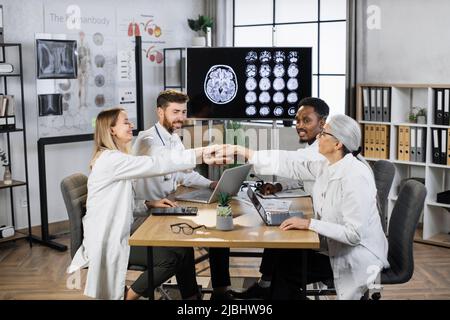 Une équipe pluriethnique souriante d'experts médicaux se réunissant les mains tout en étant assis autour d'une table avec des gadgets modernes dans la salle de réunion. Concept de réunion et de travail d'équipe réussis. Banque D'Images