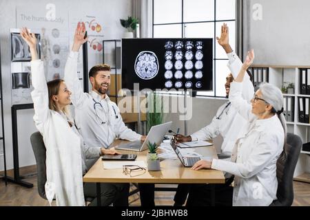 Des scientifiques internationaux motivés de divers âges assis autour d'une table dans la salle de conférence avec moniteur montrant un examen IRM du cerveau. Les médecins qui donnent des gestes forts avec leurs mains pendant la réunion. Banque D'Images