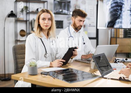 Portrait d'une femme de race blanche assise à table pendant la réunion du personnel médical avec une tablette numérique entre les mains. Médecin masculin barbu concentré travaillant sur un ordinateur portable en arrière-plan. Banque D'Images