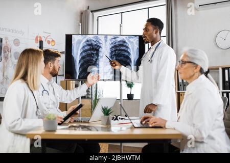 Quatre médecins multiculturels regardant l'écran numérique avec rayons X des poumons pendant la réunion du personnel médical. Un médecin afro-américain se disputant avec un collègue caucasien sur le diagnostic du patient. Banque D'Images