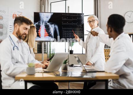 Une équipe de spécialistes médicaux compétents se réunit à la salle de conférence pour discuter de la radiographie des patients affichée sur un écran numérique moderne. Les médecins masculins et féminins coopèrent pour la réussite au travail. Banque D'Images