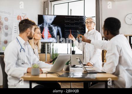 Une équipe de spécialistes médicaux compétents se réunit à la salle de conférence pour discuter de la radiographie des patients affichée sur un écran numérique moderne. Les médecins masculins et féminins coopèrent pour la réussite au travail. Banque D'Images