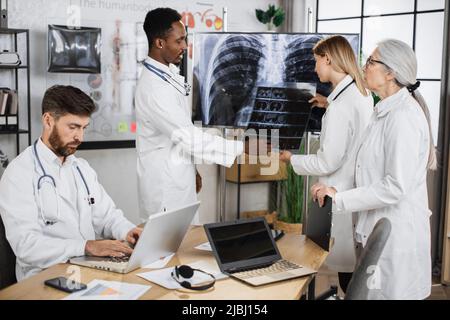 Médecin caucasien tenant un examen radiographique et parlant avec des collègues multiethniques tout en se tenant près du moniteur avec le résultat du patient. Les médecins-thérapeutes de Foure coopèrent pour établir un diagnostic. Banque D'Images