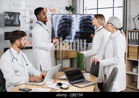 Médecin caucasien tenant un examen radiographique et parlant avec des collègues multiethniques tout en se tenant près du moniteur avec le résultat du patient. Les médecins-thérapeutes de Foure coopèrent pour établir un diagnostic. Banque D'Images