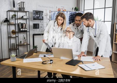 Équipe internationale de trois stagiaires ayant une réunion urgente avec une femme médecin senior dans la salle de conférence de l'hôpital. Blouses de laboratoire blanches utilisant un ordinateur portable moderne pour l'analyse du dossier médical des patients. Banque D'Images