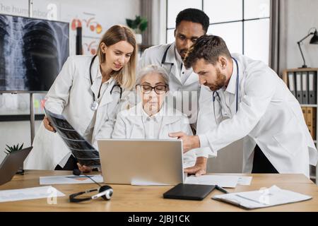 Médecins multiethniques discutant de l'acquisition de rayons X tout en se tenant autour de la femme médecin senior à l'hôpital. Femme aînée compétente assise à un bureau devant un ordinateur portable et donnant la consultation pour des collègues. Banque D'Images