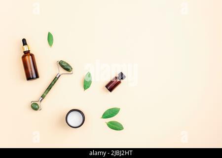 Crème dans un pot, bouteille de sérum et rouleau facial de jade sur fond beige neutre avec feuilles vertes, vue du dessus. Flat lay, espace de copie. Banque D'Images