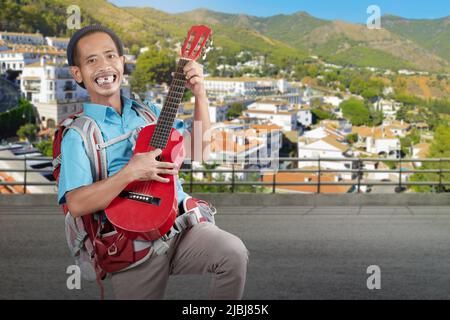 Homme asiatique avec un bonnet beanie et un sac à dos portant une guitare debout dans la rue avec un paysage urbain sur fond de colline Banque D'Images